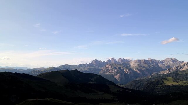 Aerial view of the rugged Sella Pass mountain range with green slopes and rocky peaks under a clear blue sky in Selva, Trentino-Alto Adige, Italy.