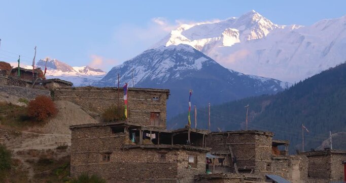 Annapurna Circuit, Manang, Nepal, drone glides toward traditional stone houses and prayer flags with snow covered Himalayan mountains in the background during late afternoon light.