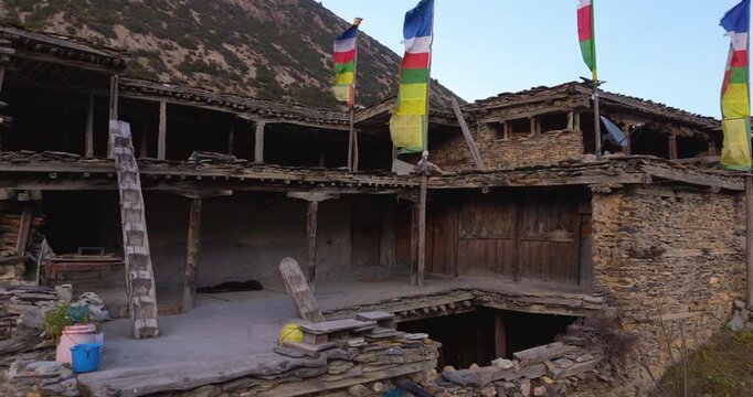 Annapurna Circuit, Manang, Nepal, drone gliding forward approaching colorful Buddhist prayer flags fluttering above traditional stone architecture in a remote mountain village.