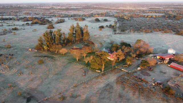 Sunrise drone shot of a remote Argentine estancia with farm buildings, livestock corrals, and water tank during a harsh winter frost