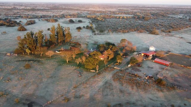 Drone shot of a remote Argentine estancia with farm buildings, livestock corrals, and water tank during a harsh winter frost