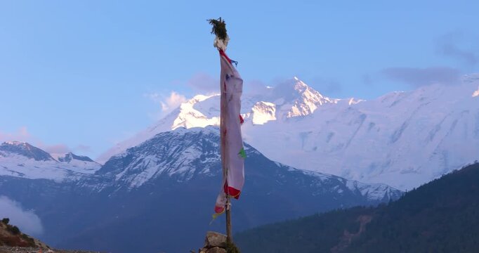 Annapurna Circuit, Manang Nepal, drone shot of a vertical Buddhist prayer flag fluttering in the wind on a hilltop with the snow-covered peak of Annapurna II in the background during evening light.