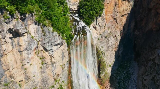 A breathtaking cinematic aerial view capturing the thunderous 106-meter drop of Boka Waterfall as turquoise glacial meltwater surges through a steep, rugged limestone canyon.