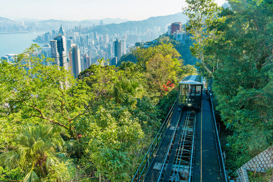 Peak tram funicular railway traveling through lush greenery with Hong Kong city skyline and Victoria harbour, China. Public transport, urban development, sustainable mobility, tourism infrastructure