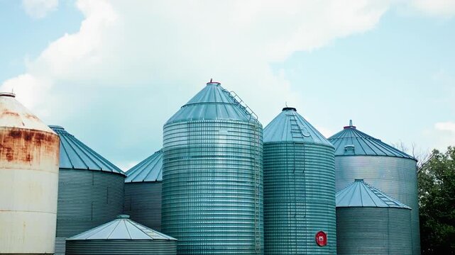 Low angle telephoto drone shot flying backward past grain silos, creating parallax movement against a bright blue summer sky