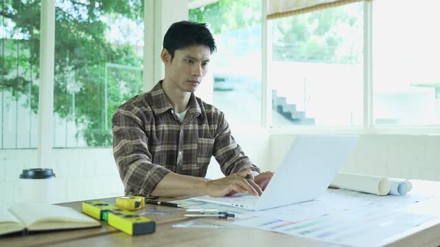 Architect working on building blueprints with a compass and laptop at a modern workspace, surrounded by rolled plans, measuring tools and color samples.