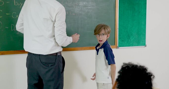 Male teacher guides young boy beside green chalkboard during math lesson, checking answer work and encouraging progress, attention, responsibility, and back to school classroom confidence daily.