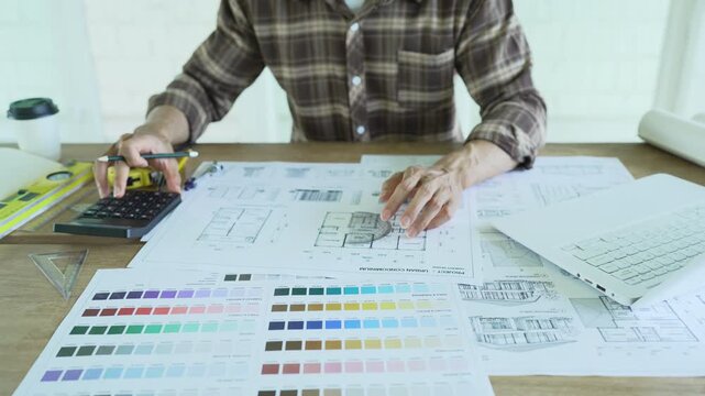 Architect drawing and reviewing detailed building blueprints on a desk with a laptop, rolled plans and color samples, representing architectural design and construction planning.