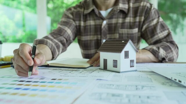 Architect drawing and reviewing detailed building blueprints on a desk with a laptop, rolled plans and color samples, representing architectural design and construction planning.