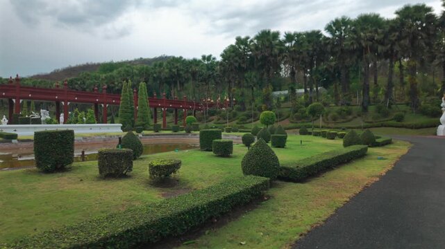 Landscaped garden park with trimmed shrubs and red bridge, Rajapruek Royal Palace lawn with topiary bushes pathway and tropical trees landscape 4k