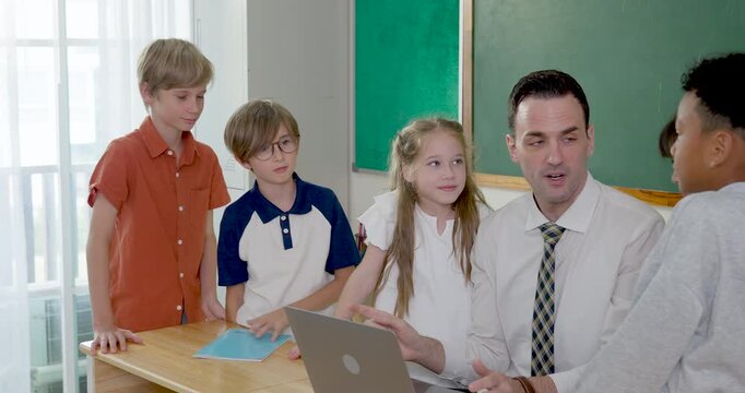 Male adult teacher uses laptop with young multicultural boy and girl student at wooden desk, guiding back to school classwork, digital learning, peer teamwork and primary study routine today
