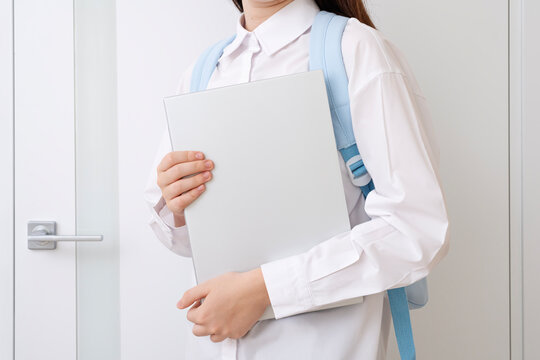 Cute teenage girl without face with backpack and notebook in hands on white interior background