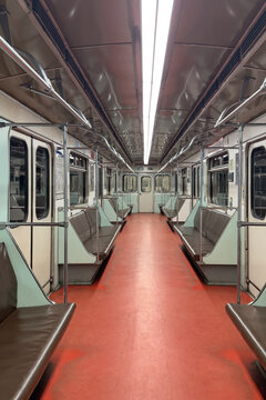 Empty subway train interior with red flooring, metal handrails, and padded benches, illuminated by overhead fluorescent lights