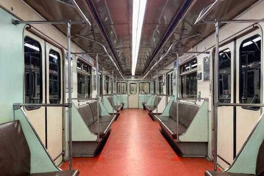 Interior view of an empty subway train featuring red flooring and green seats. Metal poles are visible throughout the space, illuminated by bright fluorescent lights above