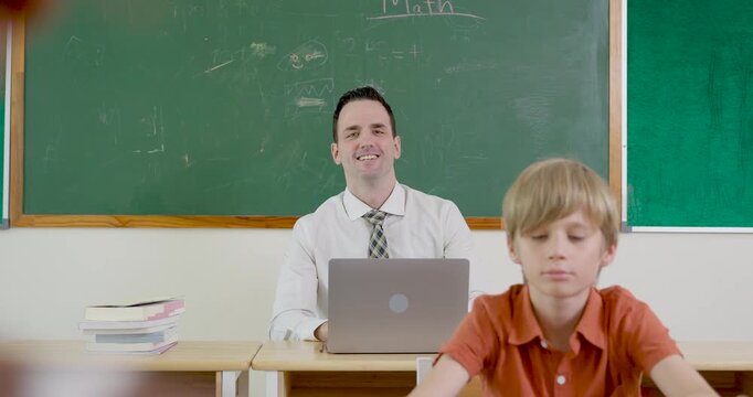 Adult male teacher works on laptop behind blond elementary boy near green chalkboard, back to school class routine builds attention, digital learning support, readiness, discipline, participation