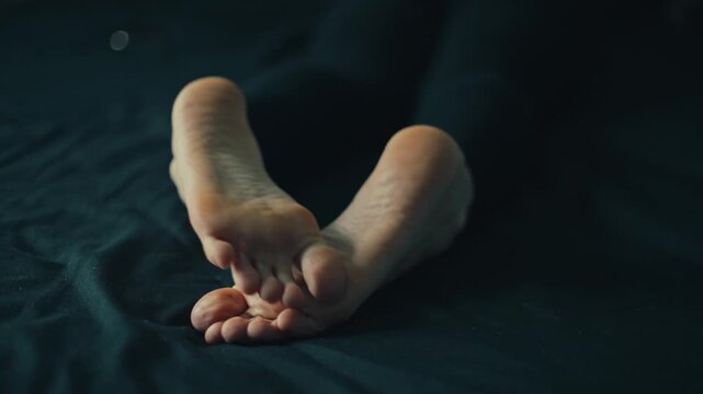 Close-up of female bare soles gently lifting and crossing on dark fabric with soft directional light revealing heel contours, toe pads, skin texture, and subtle repositioning