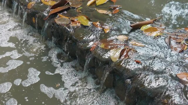 Clear water flowing over a rocky retaining wall with autumn leaves.