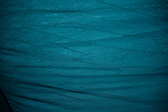 A wide close-up of water drops resting on a vibrant blue, wrinkled waterproof tent tarp. The image highlights the texture of synthetic material, folds, and hydrophobic surface during a rainy day outdo