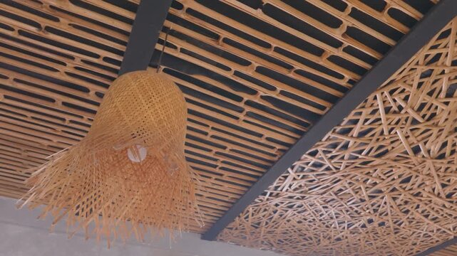 Woven bamboo lamp shade hanging from a modern decorative ceiling with wooden slats and abstract stick texture.