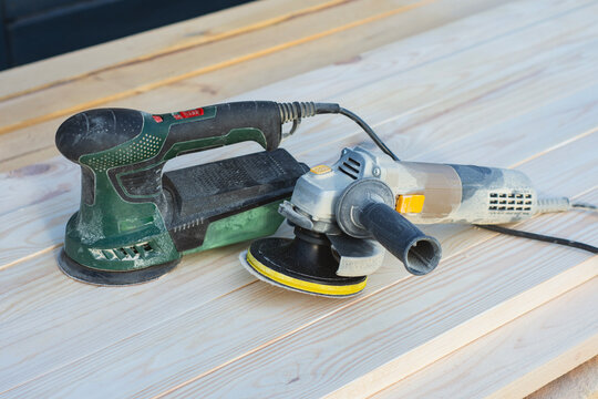 Electric orbital sander and angle grinder with sanding disc on wooden planks outdoors. Woodworking power tools, carpentry equipment or surface polishing concept for hardware store website banner.