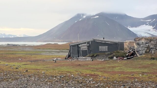 A weathered cabin endures on Svalbard's expansive Arctic tundra. Majestic, snow-dusted mountains loom beyond a tranquil fjord. Autumnal hues paint the ground under a soft, overcast sky in Norway.
