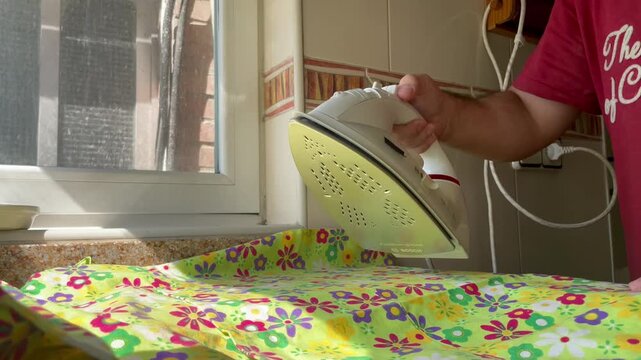 Plancha de vapor persona planchando, quitando arrugas ropa y alisar en tabla de planchar y alisando prenda con electrodom&eacute;stico agua destilada iron clothes y calor cable y enchufe en cocina y ventana