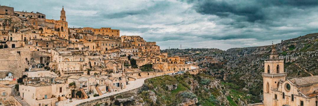 Aerial view of the historic Sassi di Matera, with stone dwellings carved into limestone cliffs, narrow streets, and a dramatic canyon under a cloudy sky in Matera, Basilicata, Italy.