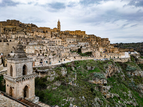 Aerial view of the historic Sassi di Matera with ancient stone dwellings, narrow streets, and a prominent bell tower under a cloudy sky in Matera, Basilicata, Italy.