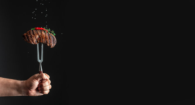Hand holding a meat fork with grilled steak garnished with herbs and spices against a dark background, showcasing culinary presentation and food preparation