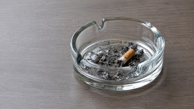 Glass ashtray holding a smoldering cigarette butt and scattered ashes on a wood tabletop