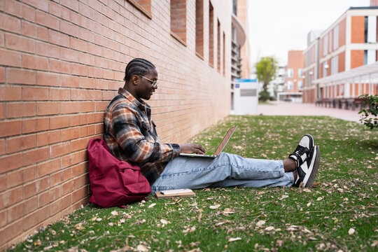 African University student with glasses working on laptop sitting on grass at Campus park