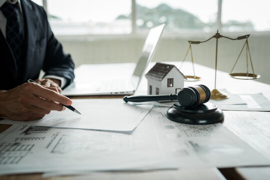 Real estate professional reviewing property documents at a desk with a house model, legal gavel, architectural plans, laptop, and scales, representing property law and transactions.