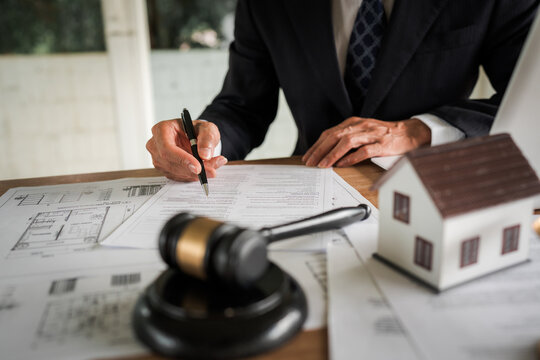 Real estate professional reviewing property documents at a desk with a house model, legal gavel, architectural plans, laptop, and scales, representing property law and transactions.