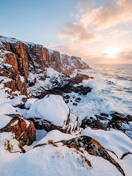 Snow covered rocky coastline with crashing ocean waves against cliffs at sunset with golden light illuminating clouds and winter landscape.