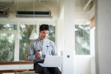 Fototapeta premium Young Asian businessman working on a laptop in a modern bright office with large windows, representing business, technology, productivity, and professional work.