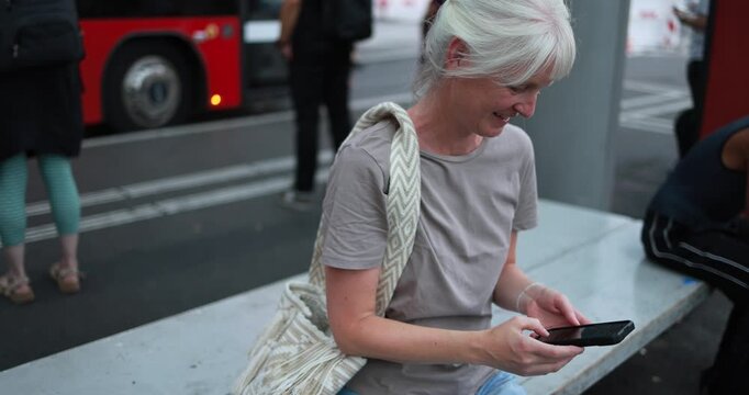 Caucasian senior woman using smartphone at bus station wearing casual clothes - City lifestyle and technology concept - UGC content