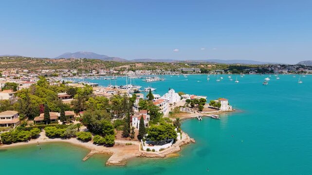 Drone reveals Porto Cheli's full peninsula in summer. Church, villas, marina and turquoise bay shine under a clear sky with mountains stretching to the horizon.