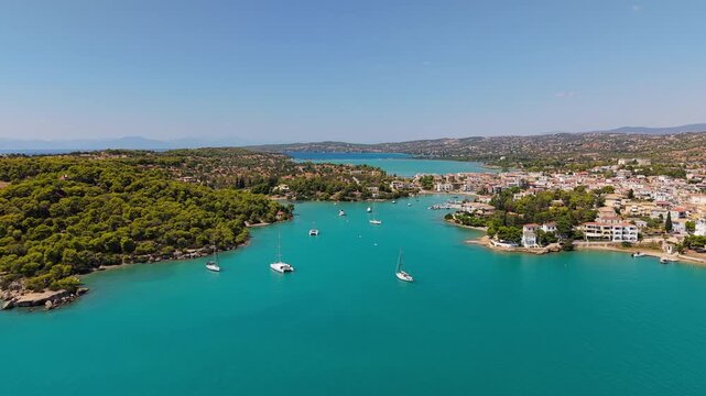 Drone flies forward over a turquoise bay toward a charming coastal village in Porto Cheli. Sailboats and catamarans drift peacefully under a clear summer sky.