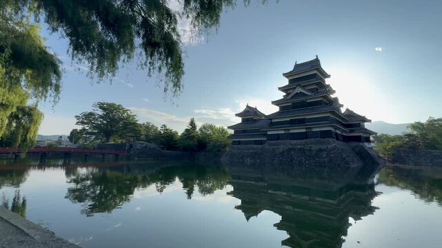 Wide static shot of historic Matsumoto Castle in Nagano Japan during a clear morning with the ancient fortress and its red bridge reflecting on the calm moat water broll