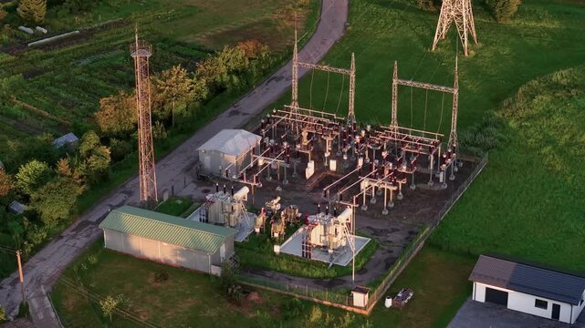 Sweeping drone video of an electrical substation surrounded by green fields and trees, captured at golden hour. Crisp, detailed aerial footage highlights industrial infrastructure in serene landscape.