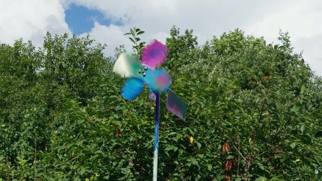 Brightly colored plastic pinwheel spins rapidly in summer breeze against lush green trees and blue sky filled with white clouds, medium shot in countryside.
