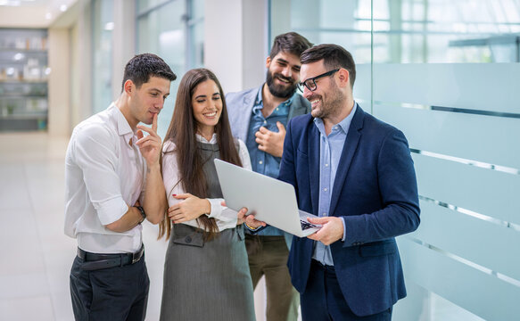 Group of happy business colleagues gathered around laptop discussing project in office hallway.