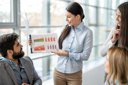 Smiling businesswoman showing clipboard with financial chart to colleagues during office meeting.