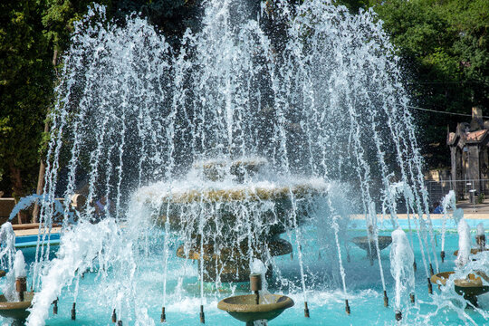 Decorative stone fountain features multiple water jets that create a misty spray in a turquoise pool, surrounded by lush greenery and trees, providing a serene outdoor atmosphere