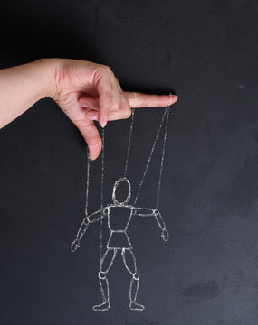 Conceptual image of manipulation, a human hand holding strings leading to a chalk-drawn puppet mannequin on a blackboard.