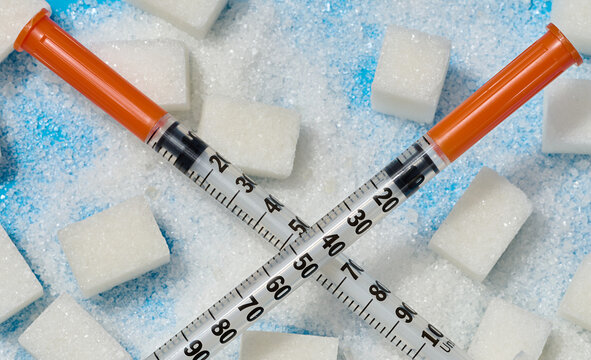 Two insulin syringes crossed over cubes and scattered white sugar on a blue background