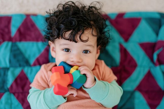 Smiling baby chewing a colorful toy on a patterned blanket for childcare banner and family wellness background design