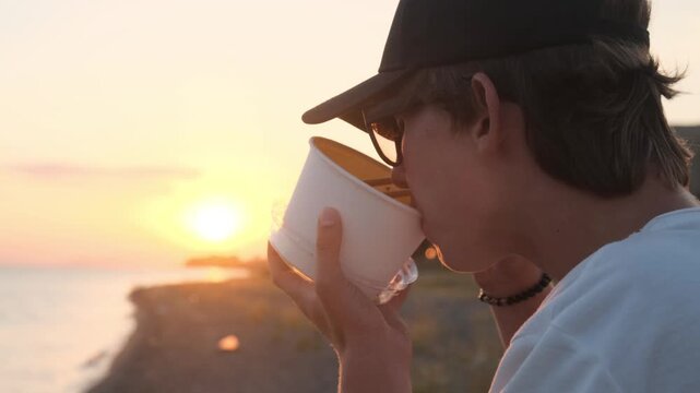 Close up, young man drinks hot instant noodle broth from a disposable container at sunset after a workout. Hot broth, beach snack, post workout meal and seaside recreation