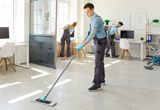 Cleaners team deliver office cleaning service. Uniformed man uses a mop on floor as coworkers dust desks and sanitize equipment in a clean modern workspace. Facility hygiene concept for firms.