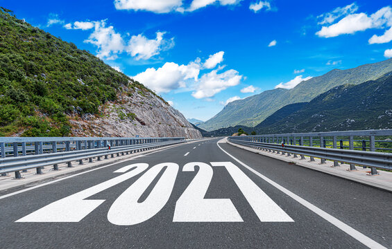 An empty highway with the year 2027 painted on the asphalt stretches towards majestic mountains under a bright blue sky.